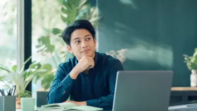 Profesional muda duduk di meja kantor sedang merefleksikan cara mengelola stres kerja dengan lebih sehat